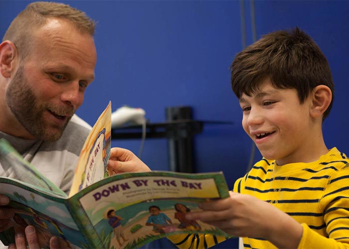 Happy, young boy smiling while reading a book with a warm, welcoming male volunteer.
